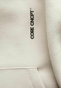 Svijetlosmeđi dukserica s kengur džepom, vidljivim šavovima i crnim logotipom "CORE CNCPNT" na gornjoj lijevoj strani. Mekana tekstura tkanine.