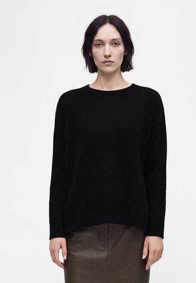 Jeune femme aux cheveux courts et foncés, portant un pull noir à manches longues et une jupe marron foncé, debout devant un fond blanc uni.
