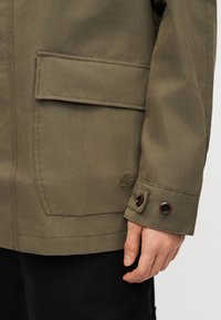 Nahaufnahme einer Hand in der Nähe der Manschette und Tasche einer olivgrünen Jacke mit Knopfdetails und einem kleinen Baum-Logo.