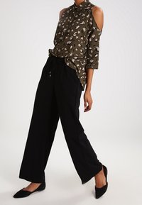 Femme portant un chemisier vert olive à épaules dénudées avec un imprimé abstrait, un pantalon noir à jambes larges et des ballerines noires pointues, marchant sur un fond blanc.