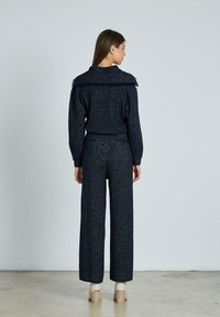 Frau steht mit dem Rücken zur Kamera, trägt ein dunkles, strukturiertes Langarmoberteil und passende weit geschnittene Hose mit hellen, hochhackigen Schuhen.