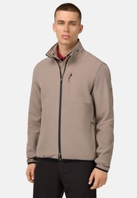 Veste softshell beige avec un col montant, fermeture éclair à l'avant, poches latérales et accents en métal noir, présentant une texture lisse et élégante.