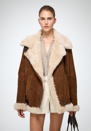 Giacca in shearling marrone con colletto beige peloso, chiusura con zip e tasche laterali. Abbinata a una camicia chiara e pantaloni corti crema.