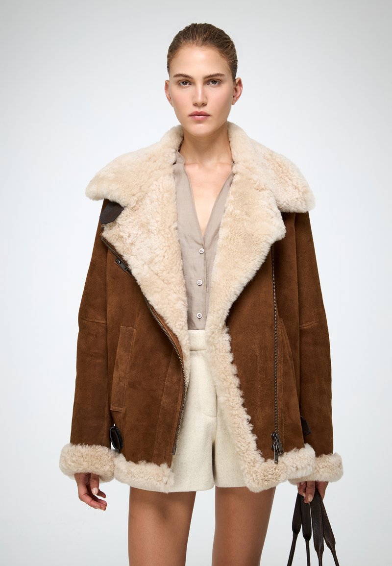 Braune Lammfelljacke mit einem flauschigen beigen Kragen, Reißverschluss und seitlichen Taschen. Mit einem hellen Hemd und cremefarbenen Shorts kombiniert.