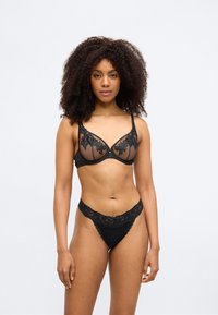 Σετ μαύρου εσωρούχου που περιλαμβάνει διαφανές σουτιέν με λεπτομέρειες από λουλουδάτη δαντέλα και αντίστοιχο στρινγκ από δαντέλα, σχεδιασμένα και τα δύο με απαλές υφές και περίπλοκα σχέδια.