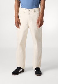 Pantalones rectos de algodón beige con un frente plano, bolsillos y presillas para cinturón. Combinados con una camiseta azul y zapatillas negras con rayas blancas.