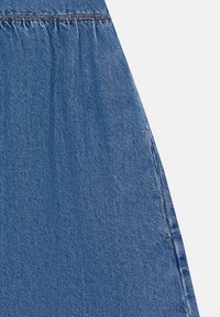 Denim tkanina v srednji modri barvi, ki ima nabran teksturo na pasu, vidne šive ob šivu in gladko površino.