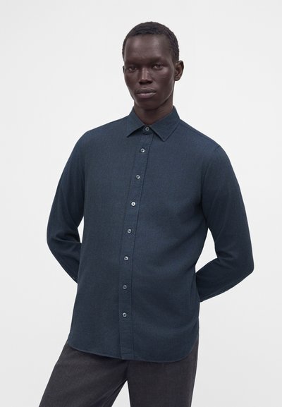 Homme portant une chemise bleu foncé boutonnée et un pantalon gris foncé, debout avec les mains derrière le dos sur un fond blanc uni.
