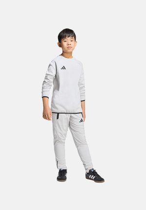 Jongen staat in een wit Adidas trainingspak en zwarte Adidas sneakers tegen een effen lichte achtergrond.