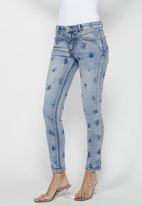 Smal blåjeans med blommig broderi i olika nyanser av blått, med en traditionell femfickorsdesign och dragkedjestängning.