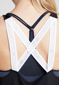 Blondyn w czarnej i granatowej bluzie z białymi elastycznymi paskami w krzyż na plecach, oznaczonymi napisem "HERO BABYLON".