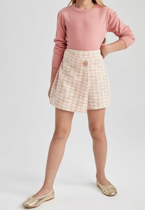 Dziewczyna w różowym swetrze z długim rękawem, beżowej, kratkowanej spódnicy z guzikiem i złotych balerinach, stoi z jedną ręką na biodrze.