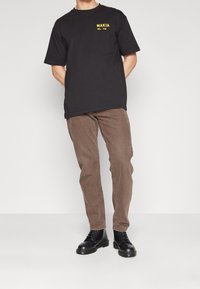 Mies pukeutuneena mustaan "MAKIA"-T-paitaan, ruskeisiin farkkuihin ja mustiin nauhallisiin saappaisiin, seisoo valkoisella taustalla kädet selän takana.