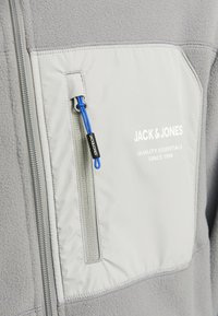 Chaqueta de forro polar gris con un tejido texturizado, un bolsillo en el pecho con cremallera y un tirador azul, y un logo de marca impreso en blanco.