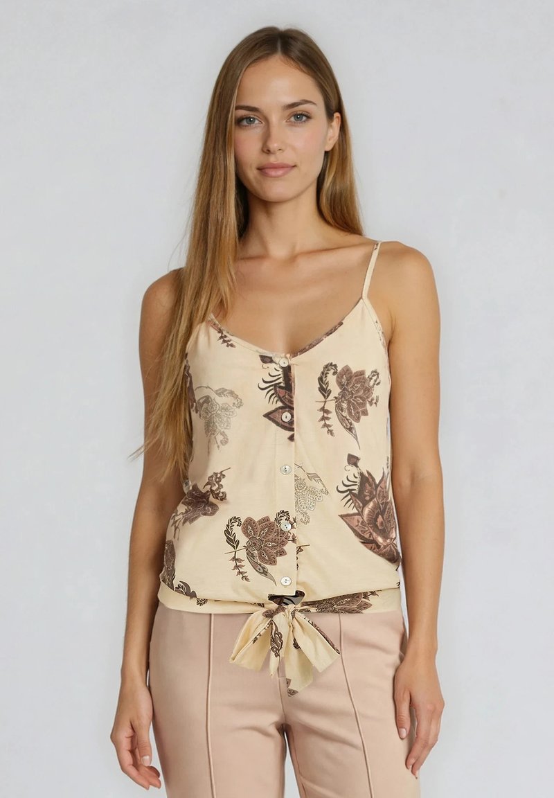 Femme portant un haut sans manches beige à fleurs boutonné, noué à la taille, avec un pantalon rose clair, debout devant un fond uni.
