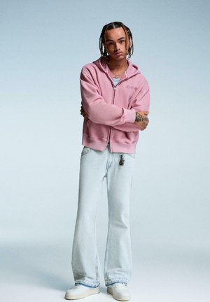 Giovane con capelli intrecciati che indossa una felpa con cappuccio rosa, jeans azzurri chiari con accessorio a catena e scarpe da ginnastica bianche, in piedi davanti a uno sfondo semplice.