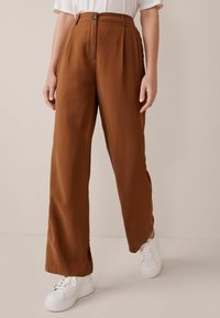 Braune, weit geschnittene Hosen aus glattem Stoff mit vorderem Knopfverschluss. Kombiniert mit weißen Sneakers auf einem neutralen Hintergrund.