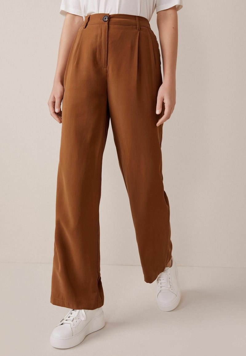 Braune, weit geschnittene Hosen aus glattem Stoff mit vorderem Knopfverschluss. Kombiniert mit weißen Sneakers auf einem neutralen Hintergrund.