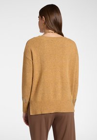 Maglione oversize giallo senape con orlo a costine, caratterizzato da borchie decorative sui polsini e un tessuto testurizzato.
