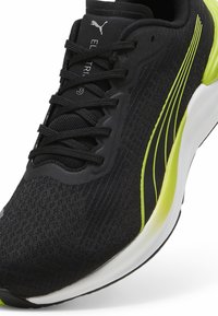 Zapatilla de correr negra con parte superior de malla texturizada, acentos amarillos brillantes y puntera sintética. Presenta un sistema de cordones y una suela blanca acolchada.