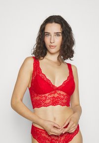 Červený čipkovaný bralet s hlbokým výstrihom a zúbkovanými okrajmi, s priesvitným čipkovaným pásom, ktorý zvýrazňuje priliehavý siluetu.
