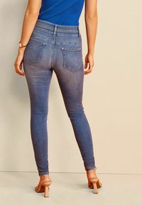 Jeans de mezclilla azul delgados con una cintura alta, que cuentan con dos bolsillos traseros y un sutil efecto de desgaste. Complementados con tacones de tiras beige.