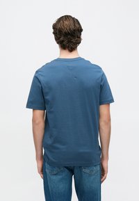 Μπλε βαμβακερό μπλουζ άκι με κοντά μανίκια, στρογγυλή λαιμόκοψη και μικρή τρίγωνη λεπτομέρεια στο πίσω μέρος. Η υφή είναι απαλή και καθημερινή.