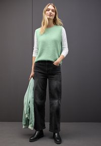 Miętowy polar z długim rękawem, założony na białą koszulę z długim rękawem, w połączeniu z czarnymi spodniami o szerokich nogawkach i czarnymi botkami za kostkę. Trzyma dopasowaną kurtkę.