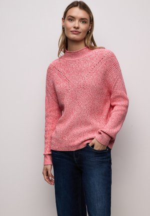 Femme portant un pull en tricot rose avec des détails à motifs et un jean bleu, debout avec une main dans la poche devant un fond uni.