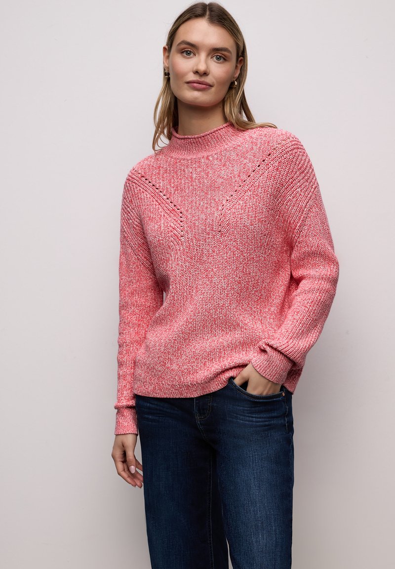 Femme portant un pull en tricot rose avec des détails à motifs et un jean bleu, debout avec une main dans la poche devant un fond uni.