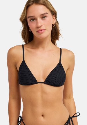 Młoda kobieta w czarnym, teksturowanym, trójkątnym topie bikini z cienkimi ramiączkami i złotymi kolczykami-kołami na tle jednolitego jasnego tła.