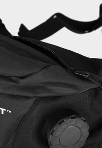Black fabric bag with a zip closure, textured surface, and a circular black hardware piece at the bottom. Logo visible on one side.