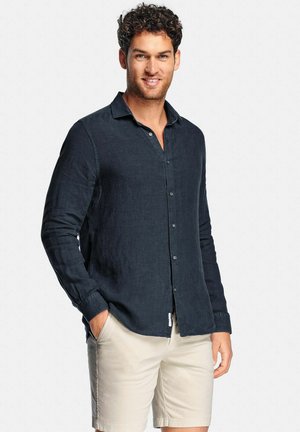 Homme portant une chemise noire à manches longues boutonnée et un short beige, debout avec une main dans la poche, souriant devant un fond uni.