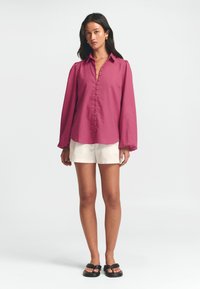 Long-sleeve, button-up shirt in magenta fabric with subtle texture, featuring a collar and puffed sleeves, paired with white shorts and black sandals.