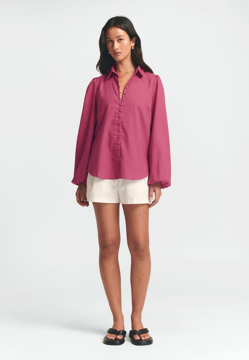 Long-sleeve, button-up shirt in magenta fabric with subtle texture, featuring a collar and puffed sleeves, paired with white shorts and black sandals.
