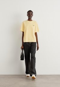 T-shirt en coton jaune à manches courtes, associé à un pantalon gris foncé et des baskets blanches. Le mannequin tient un petit sac à main noir.
