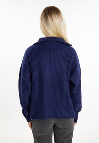 Marineblauwe geribde trui met een sjawl kraag en brede mouwen, voorzien van een textuur gebreid patroon en een ontspannen pasvorm, gecombineerd met grijze broek.