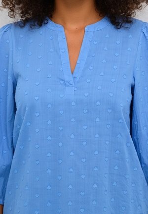 Blauwe blouse met getextureerde hartpatronen, lange mouwen en een V-hals gedragen door een persoon met krullend haar.