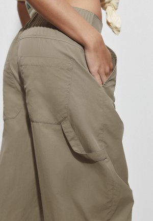 Közeli kép egy személyről, aki laza bézs cargo nadrágot visel, kezét elöl lévő zsebbe helyezve, bemutatva a zseb és a szövet részleteit.