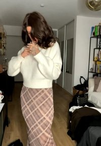Person mit langen dunklen Haaren, die einen weißen Pullover richtet, trägt einen rosa-beigen karierten Rock und steht in einem Raum mit Bücherregalen und Couch.