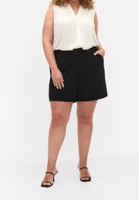 Kvinna som bär en ärmlös vit blus, svarta höga shorts med knappar och svarta klacksandaler, stående med ena handen i fickan.