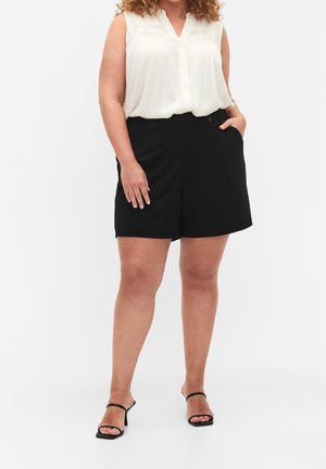 Žena v bezrukávovém bílém topu, černých šortkách s vysokým pasem a knoflíky, a černých sandálech na podpatku, stojící s jednou rukou v kapse.