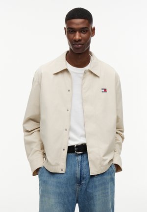 Νέος άνδρας φορώντας μπεζ σακάκι με κουμπιά πάνω από λευκό πουκάμισο και μπλε τζιν, στέκεται με τα χέρια στις τσέπες του μπροστά από λευκό φόντο.