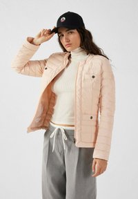 Giacca imbottita rosa chiaro con zip anteriori, due tasche sul petto e un'etichetta rotonda con logo. Collo alto a coste bianco e pantaloni grigi con cordino.