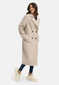 Beige wollen blend met dubbele rij knopen, zwarte knopen, zijzakken en een reverskraag, gecombineerd met blauwe spijkerbroeken en lichtgekleurde sneakers.