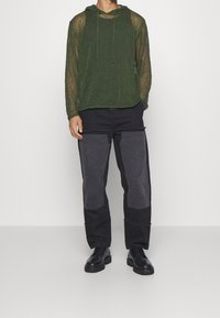 Felpa verde trasparente a maniche lunghe con cappuccio e lacci, abbinata a pantaloni neri con dettagli grigi a toppa e taglio dritto, completata da scarpe nere.