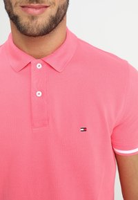 Homme portant un polo rose vif avec des boutons blancs et un petit logo rectangulaire sur le côté gauche de la poitrine, sur un fond uni.