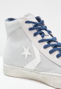 Vysoké tenisky vyrobené ze světle šedé plátěné látky s modrými tkaničkami, bílým hvězdicovým logem a gumovou podrážkou. Obsahují šité detaily a polstrovaný límec.