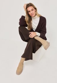 Béžové kotníkové boty s robustní podrážkou. Modelka má na sobě hnědý kardigán a široké hnědé kalhoty, sedí před světlým pozadím.