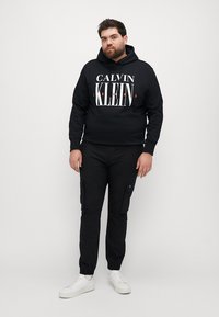 Hanorac negru cu logo-ul "CALVIN KLEIN JEANS" în alb și roșu. Asortat cu pantaloni cargo negri și adidași albi. Outfit casual.